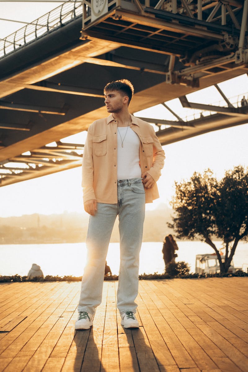 Stylish young man in casual attire poses under a bridge with a sunlit backdrop in Türkiye.